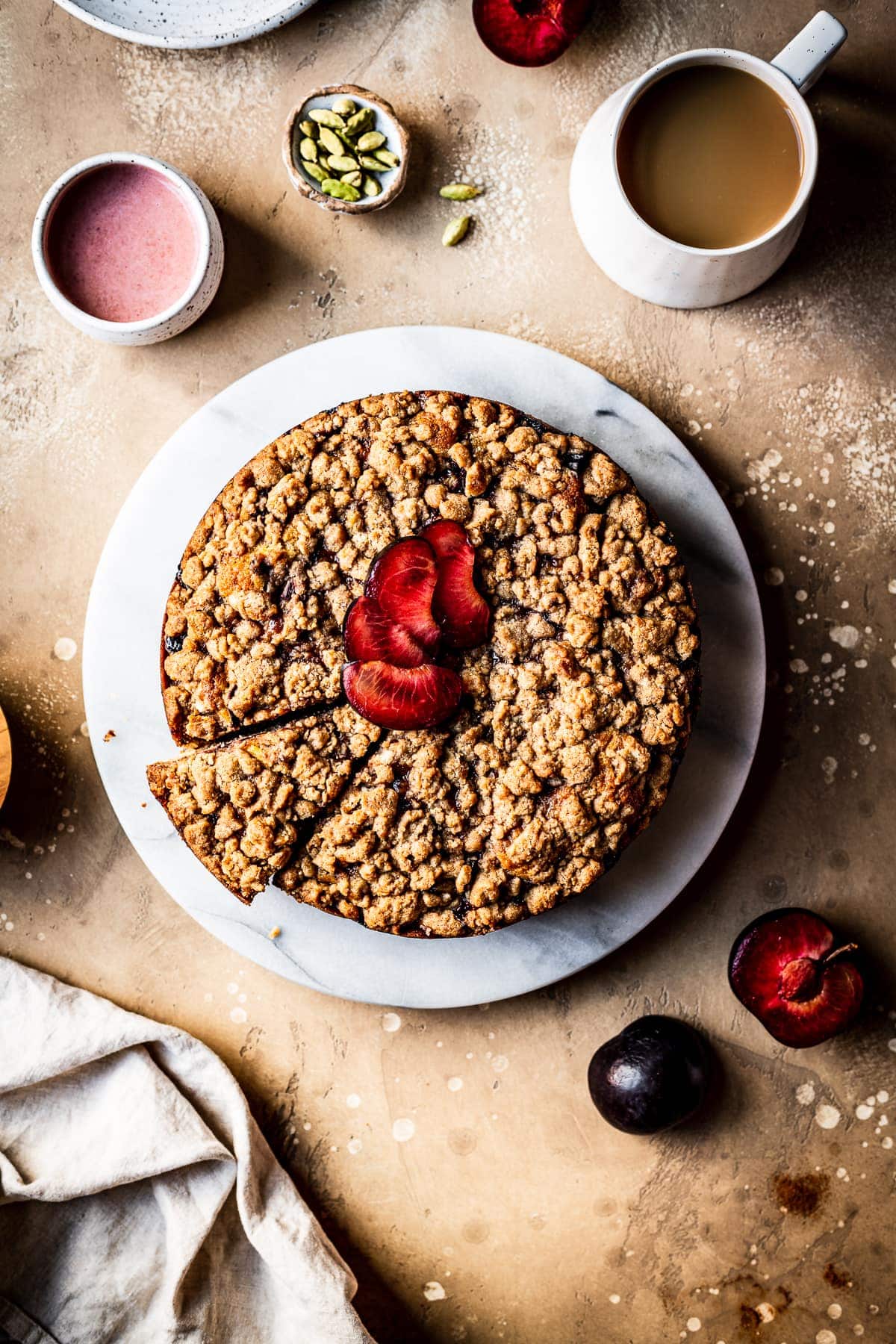 A round cake with a slice cut out of it on a marble platter on a warm tan surface. There is a fan of sliced plums on top of the cake. Nearby are a container of pink glaze, several forks, a natural linen napkin, a white ceramic coffee cup filled with coffee, a few plums sliced in half, and a small bowl of green cardamom pods.