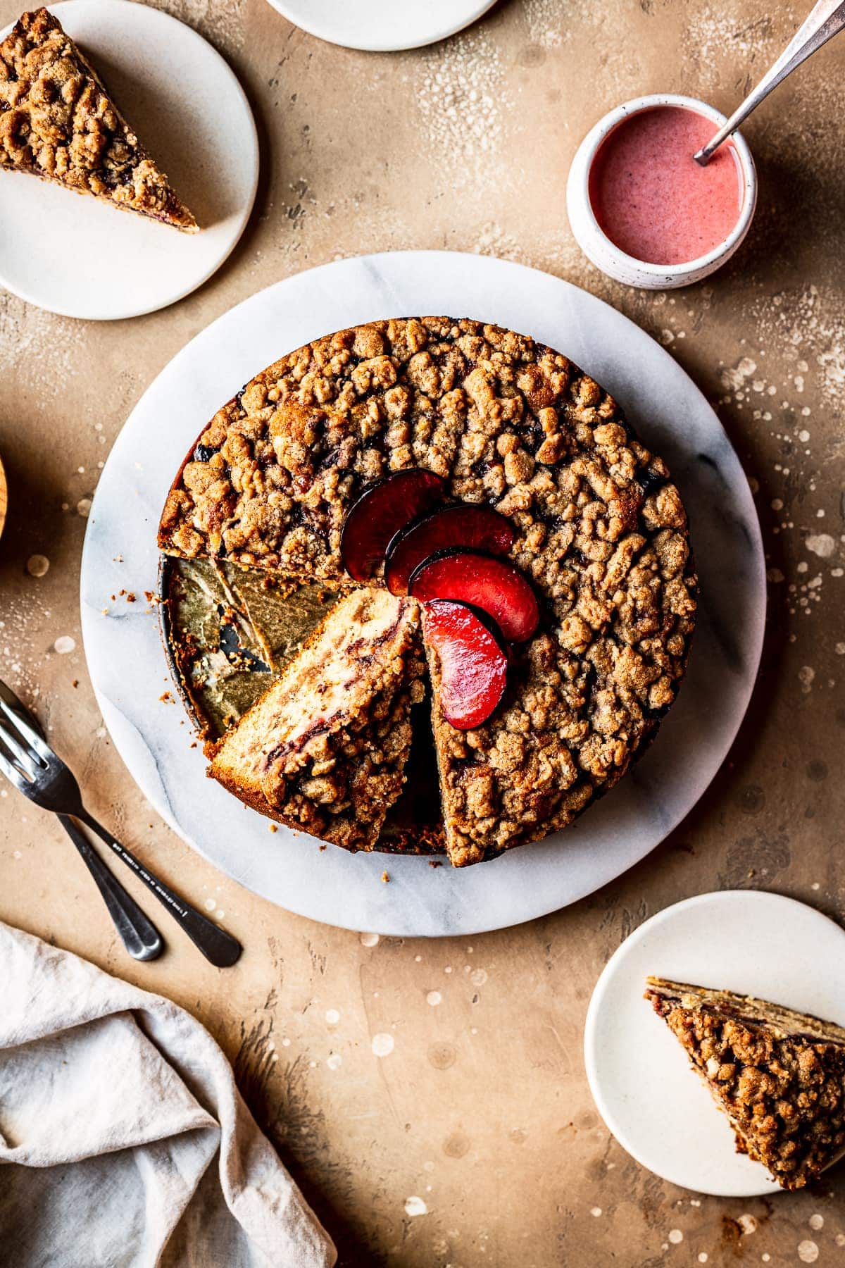 A round cake with several slices cut out of it on a marble platter on a warm tan surface. There is a fan of sliced plums on top of the cake. Nearby are several slices on small plates, a container of pink glaze, several forks and a natural linen napkin.