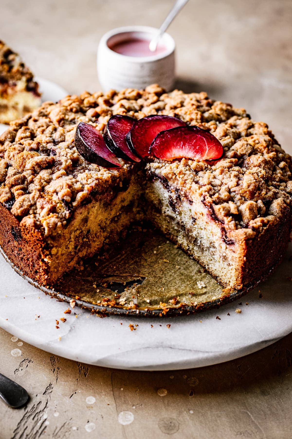 A round coffee cake with several slices cut out of it on a marble platter on a warm tan surface. There is a fan of sliced plums on top of the cake. In the background is a slice of cake and a white ceramic container of pink glaze.