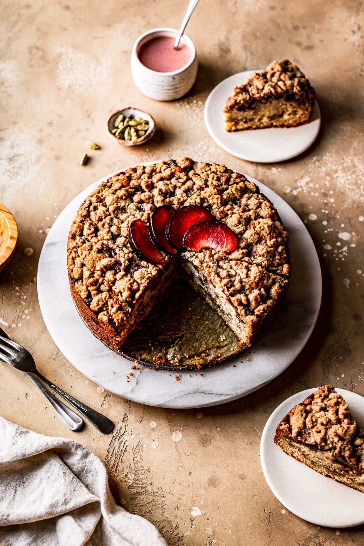 A round cake with several slices cut out of it on a marble platter on a warm tan surface. There is a fan of sliced plums on top of the cake. Nearby are several slices on small plates, a container of pink glaze, several forks, a natural linen napkin, and a small bowl of green cardamom pods.