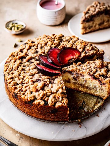 A round coffee cake with several slices cut out of it on a marble platter on a warm tan surface. There is a fan of sliced plums on top of the cake. Nearby are several slices on small plates, a container of pink glaze, and a small bowl of green cardamom pods.