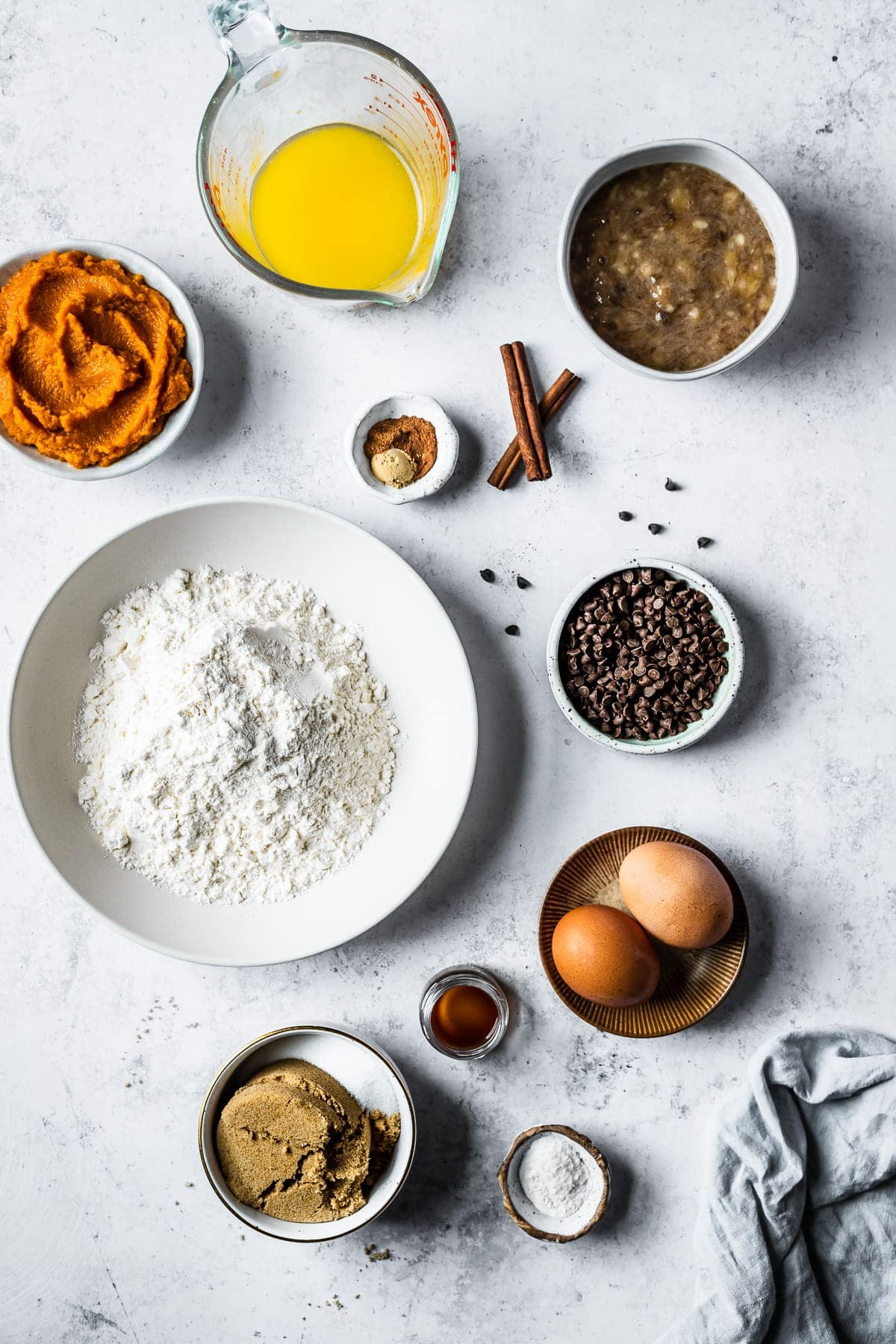 Ingredients in bowls arranged on a white textured stone surface. Ingredients include flour, pumpkin puree, melted butter, mashed bananas, eggs, sugar and spices.