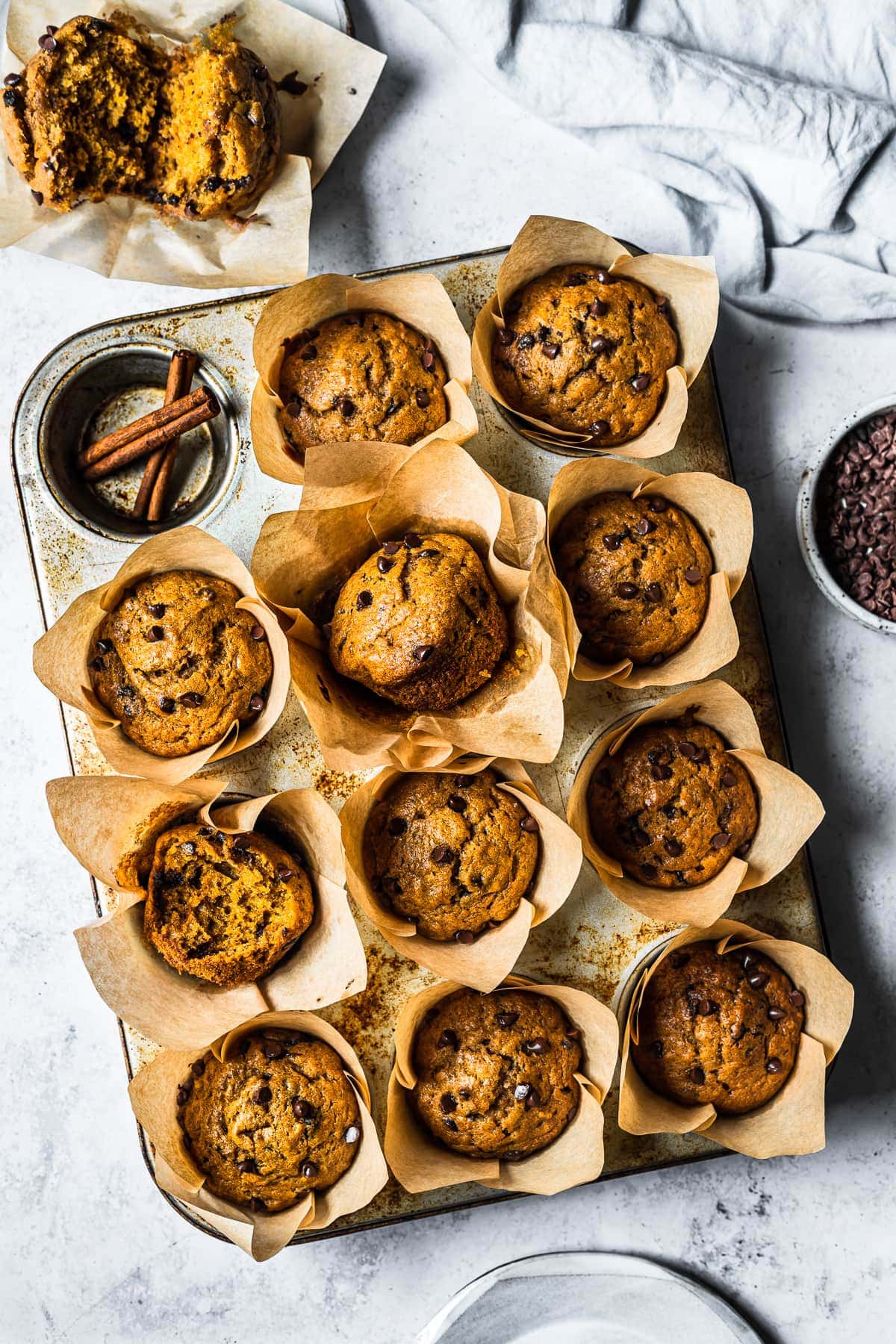 Baked pumpkin banana muffins in brown parchment liners in a muffin tin on a white stone surface. Surrounded by an opened muffin, a blue linen, a bowl of chocolate chips and a ceramic plate.