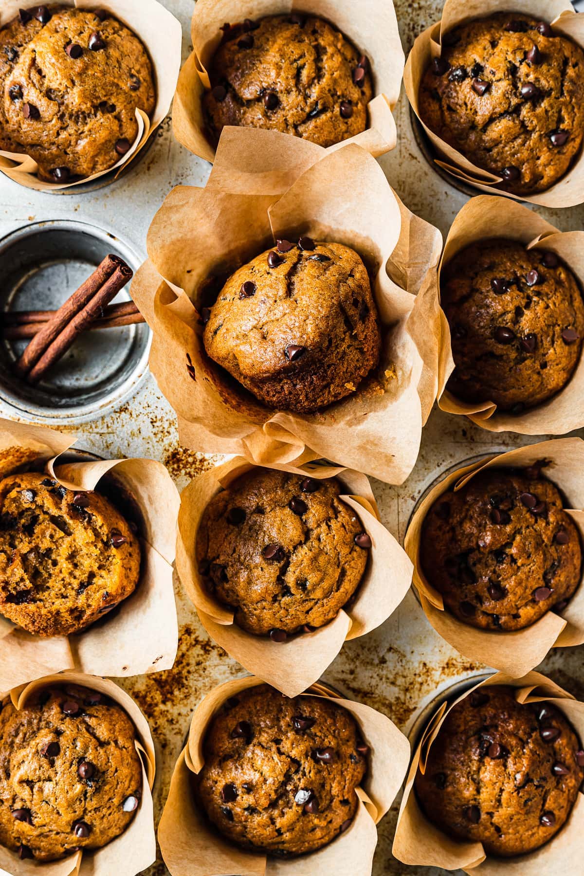 Baked muffins in brown parchment liners in a muffin tin. One cavity is empty and holds two cinnamon sticks.