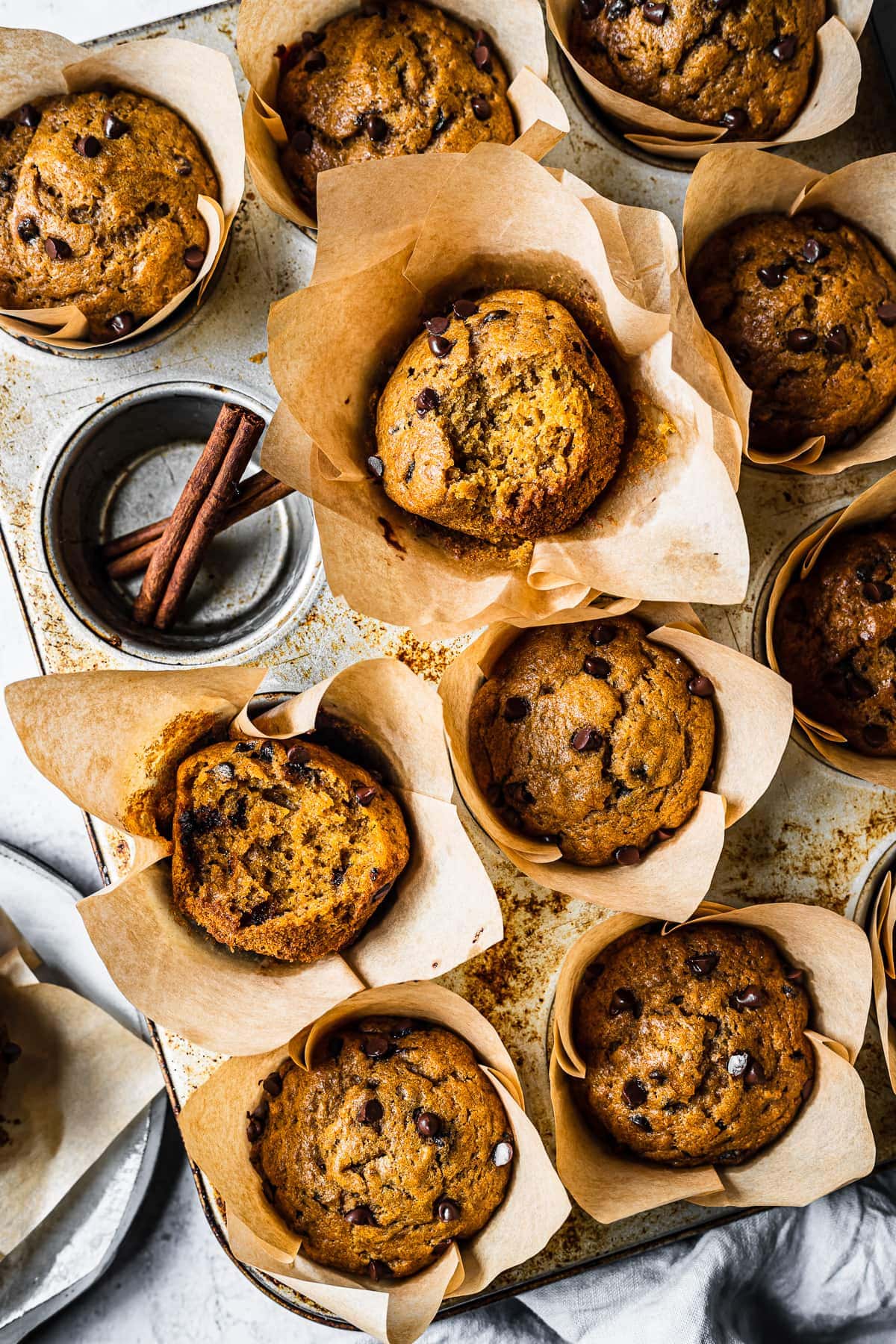 Baked pumpkin banana muffins in brown parchment liners in a muffin tin. One cavity is empty and holds two cinnamon sticks. The pan is on an angle and there are bites out of two muffins, revealing the inside texture.