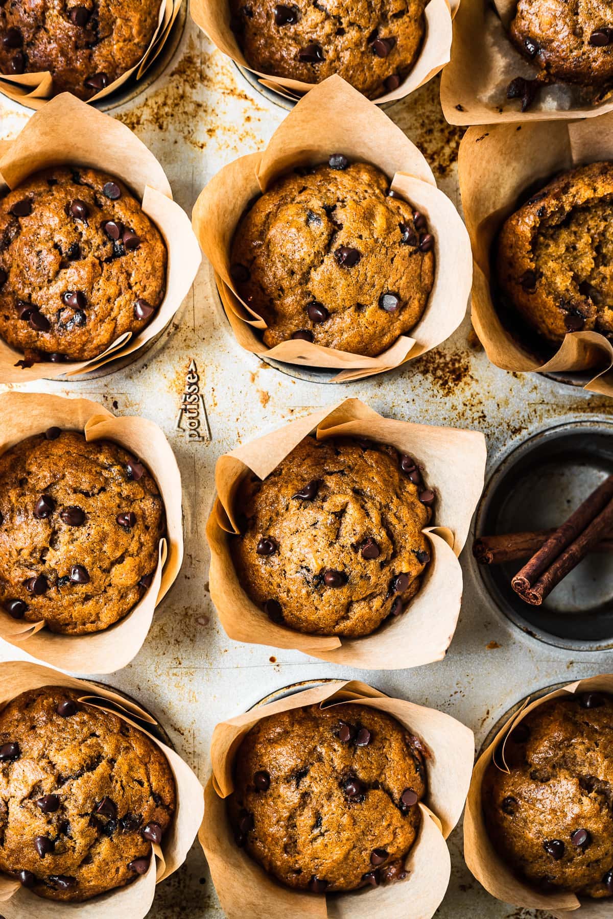 Baked muffins in brown parchment liners in a muffin tin. One cavity is empty and holds two cinnamon sticks.