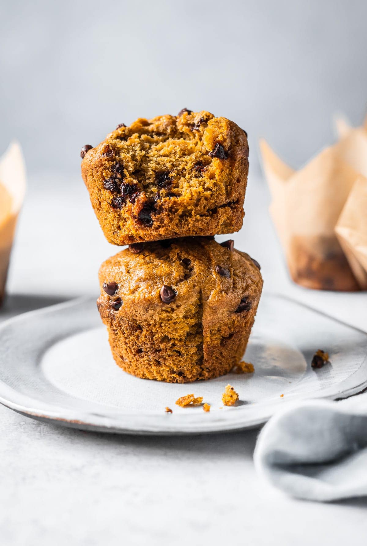 Two muffins stacked on top of each other on a white ceramic plate resting on a white stone surface. More muffins in parchment paper liners peek out from each side of the frame.