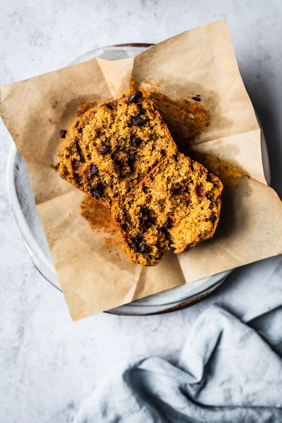 A muffin is split in half, resting face up on a brown parchment liner. It sits on a white ceramic plate on a white textured surface. A light blue linen napkin is nearby.