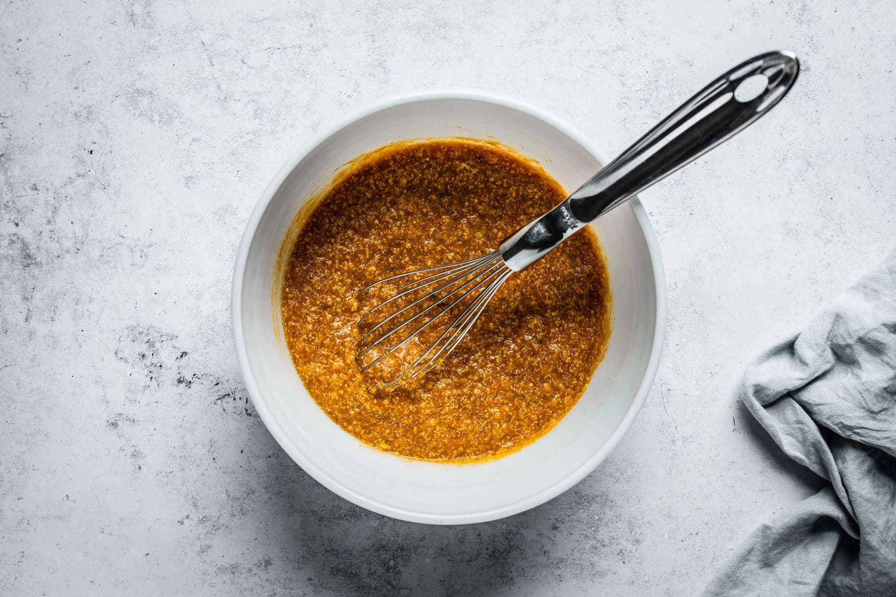 Process shot of muffin batter in a white glass mixing bowl on a textured white stone background with wet ingredients mixed together.