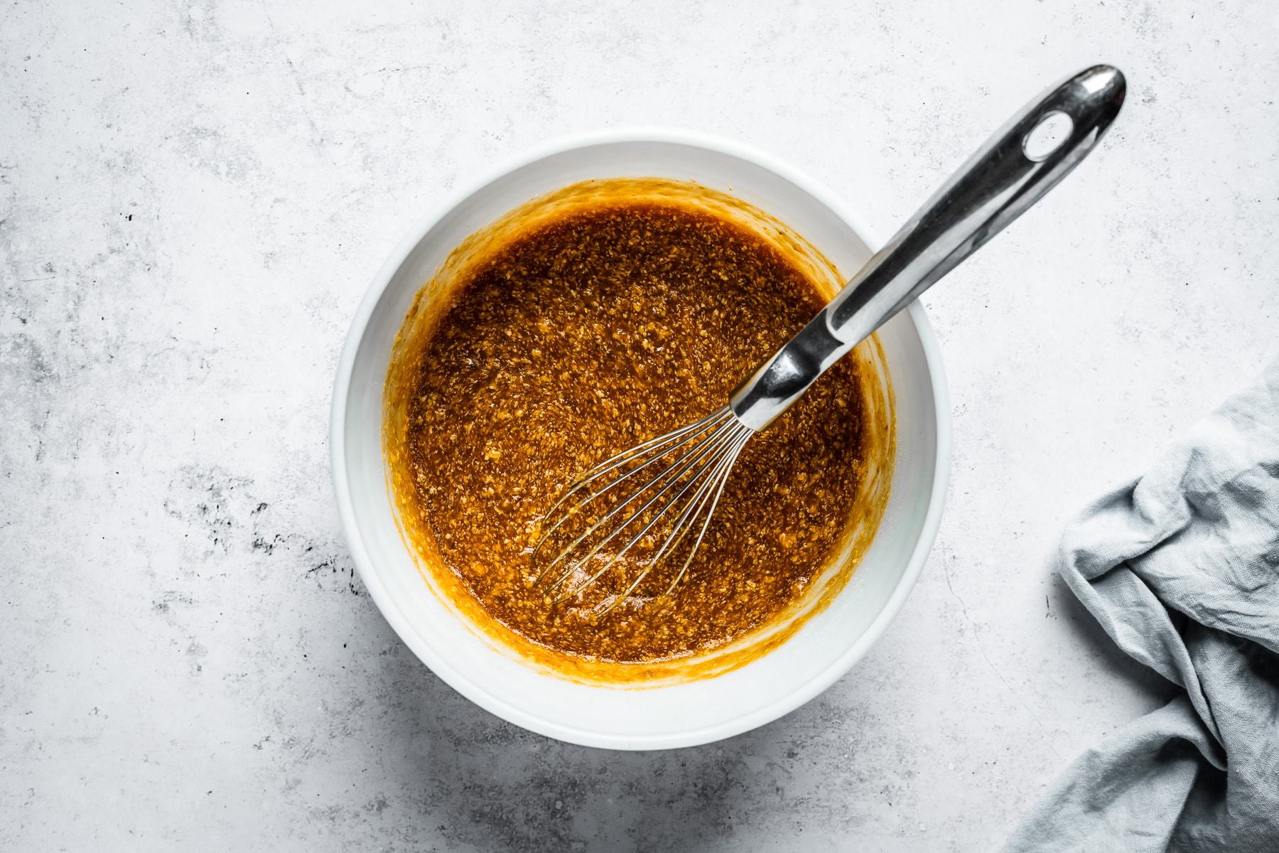 Process shot of muffin batter in a white glass mixing bowl on a textured white stone background with sugars mixed in.