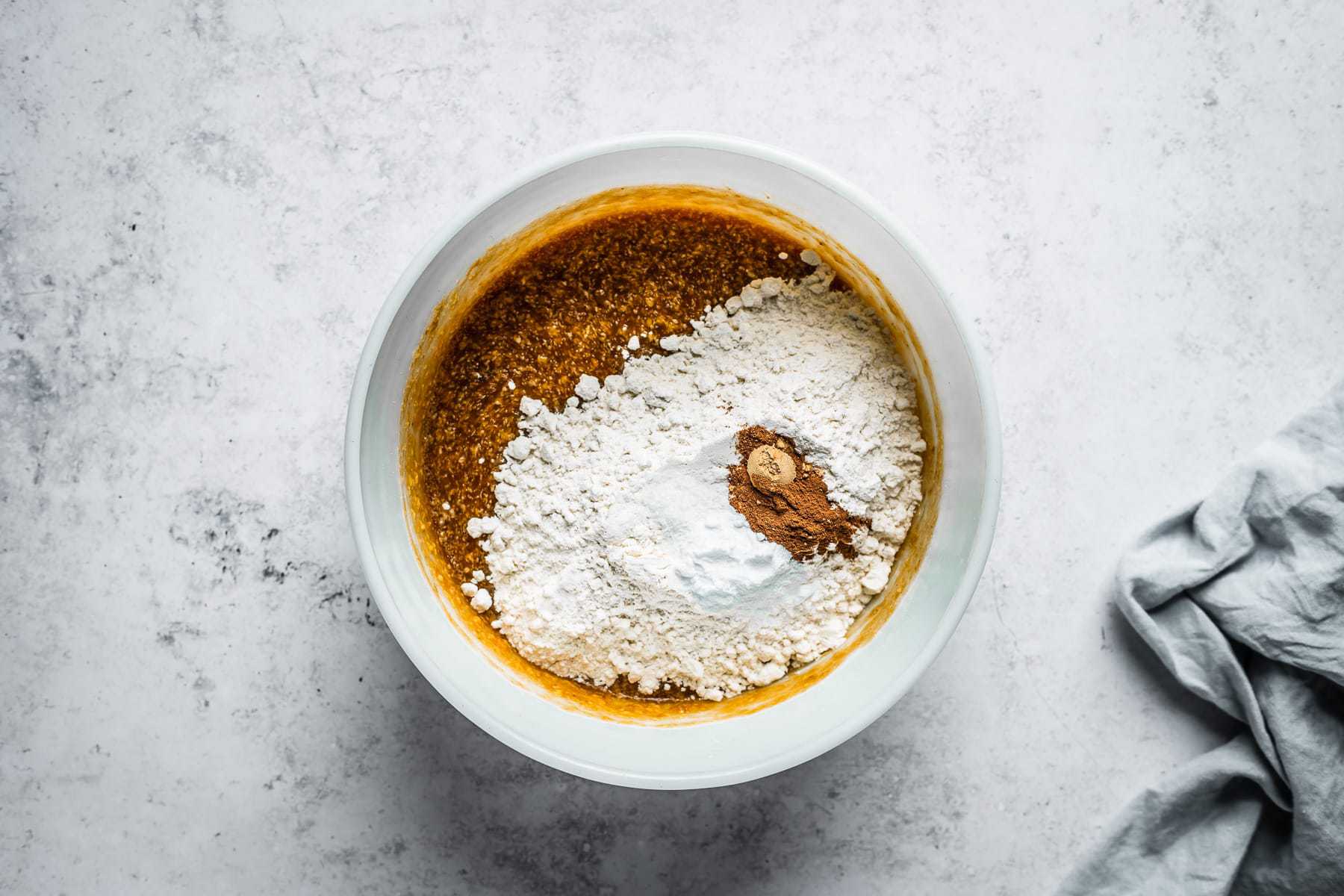 Process shot of muffin batter in a white glass mixing bowl on a textured white stone background with dry ingredients added.