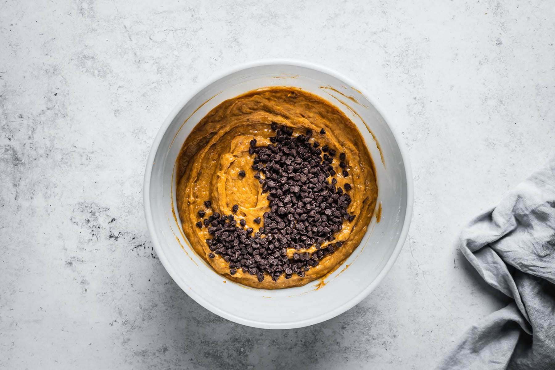 Process shot of muffin batter in a white glass mixing bowl on a textured white stone background with mini chocolate chips added.