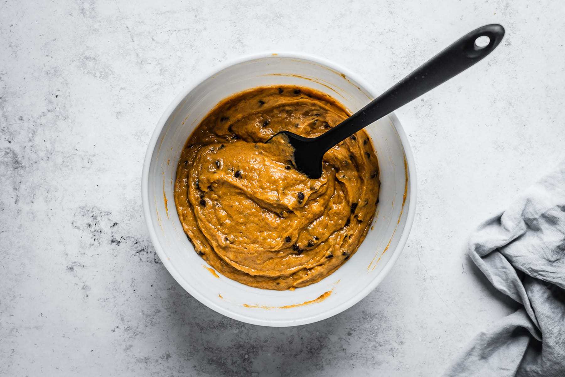 Process shot of finished muffin batter in a white glass mixing bowl on a textured white stone background.