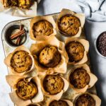 Baked pumpkin banana muffins in brown parchment liners in a muffin tin on a white stone surface. Surrounded by an opened muffin, a blue linen, a d a small ceramic bowl of chocolate chips.