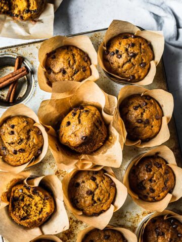 Baked pumpkin banana muffins in brown parchment liners in a muffin tin on a white stone surface. Surrounded by an opened muffin, a blue linen, a d a small ceramic bowl of chocolate chips.