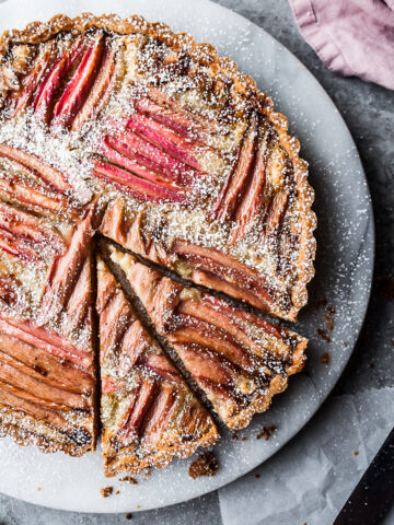 A pink hued tart dusted with powdered sugar, with two slices cut out. The tart rests on a marble serving platter on a grey stone surface. A pink napkin peeks into the frame at top right.
