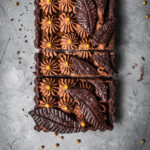 A rectangular chocolate tart with piped chocolate over layers of caramel in a chocolate pastry shell. There are decorative chocolate pastry leaves on top. It is resting on a grey textured surface.