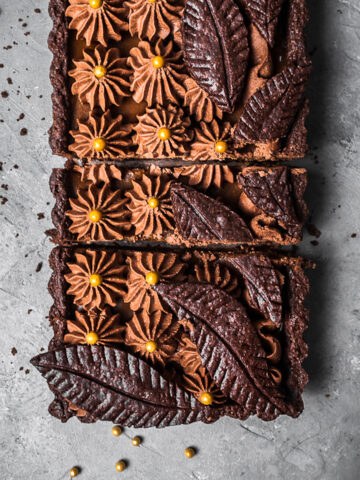 A rectangular chocolate tart with piped chocolate over layers of caramel in a chocolate pastry shell. There are decorative chocolate pastry leaves on top. It is resting on a grey textured surface.