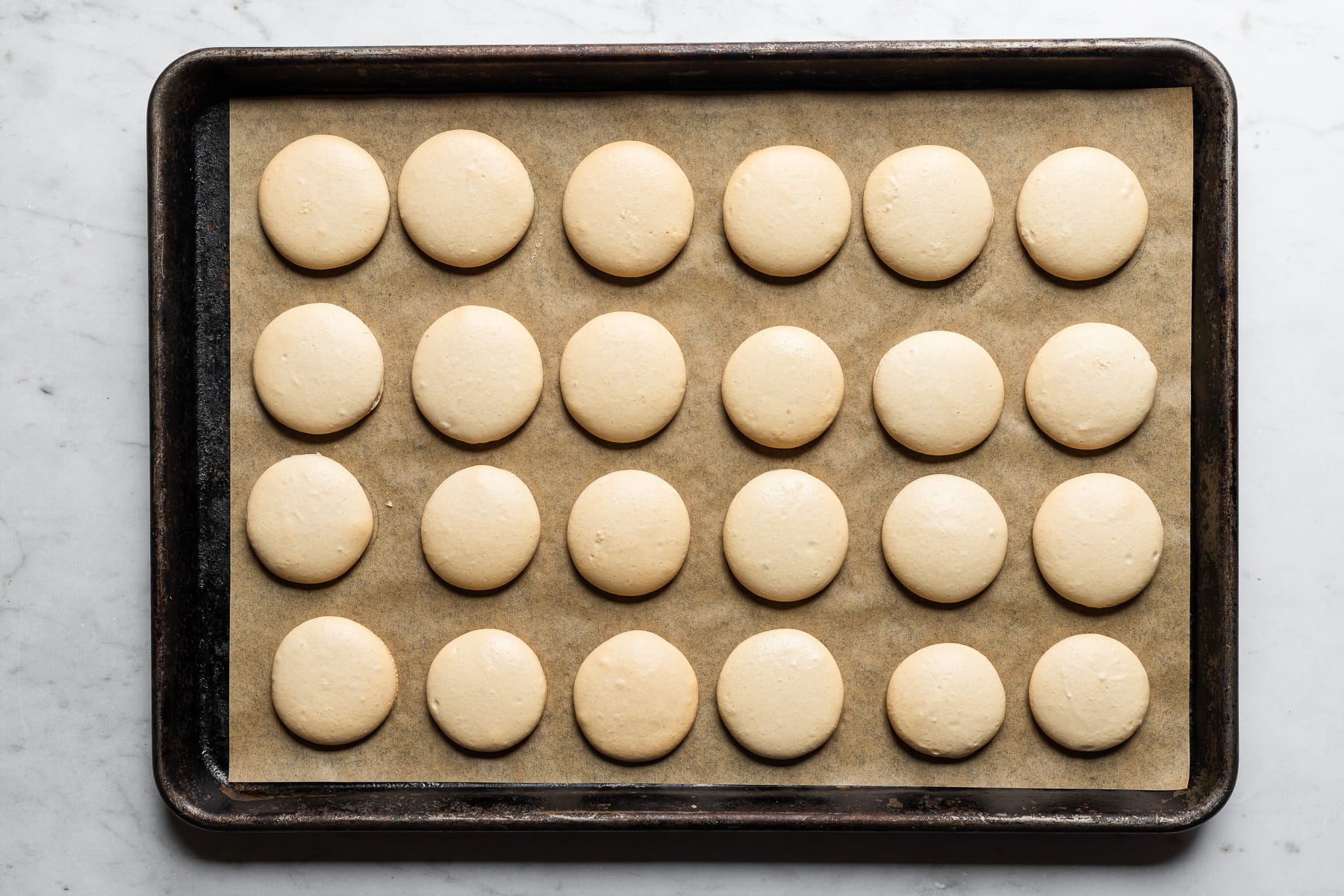 A process photo showing baked macaron shells on a parchment lined baking sheet. The baking sheet rests on a white marble background.
