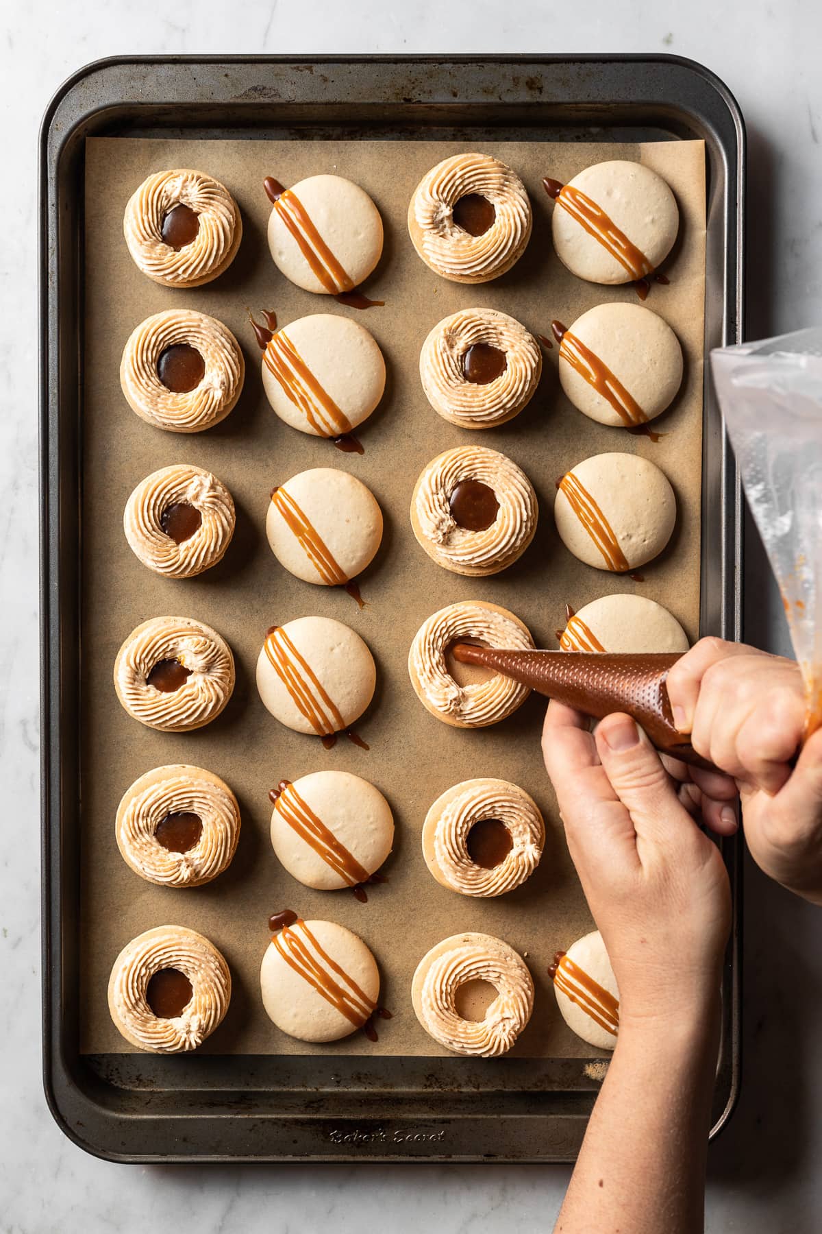 Process photo showing a parchment lined baking sheet full of macaron shells. Half are filled with buttercream and caramel sauce, and the other half have a decorative caramel decoration on top. A pair of hands holding a caramel filled piping bag reach into the frame.