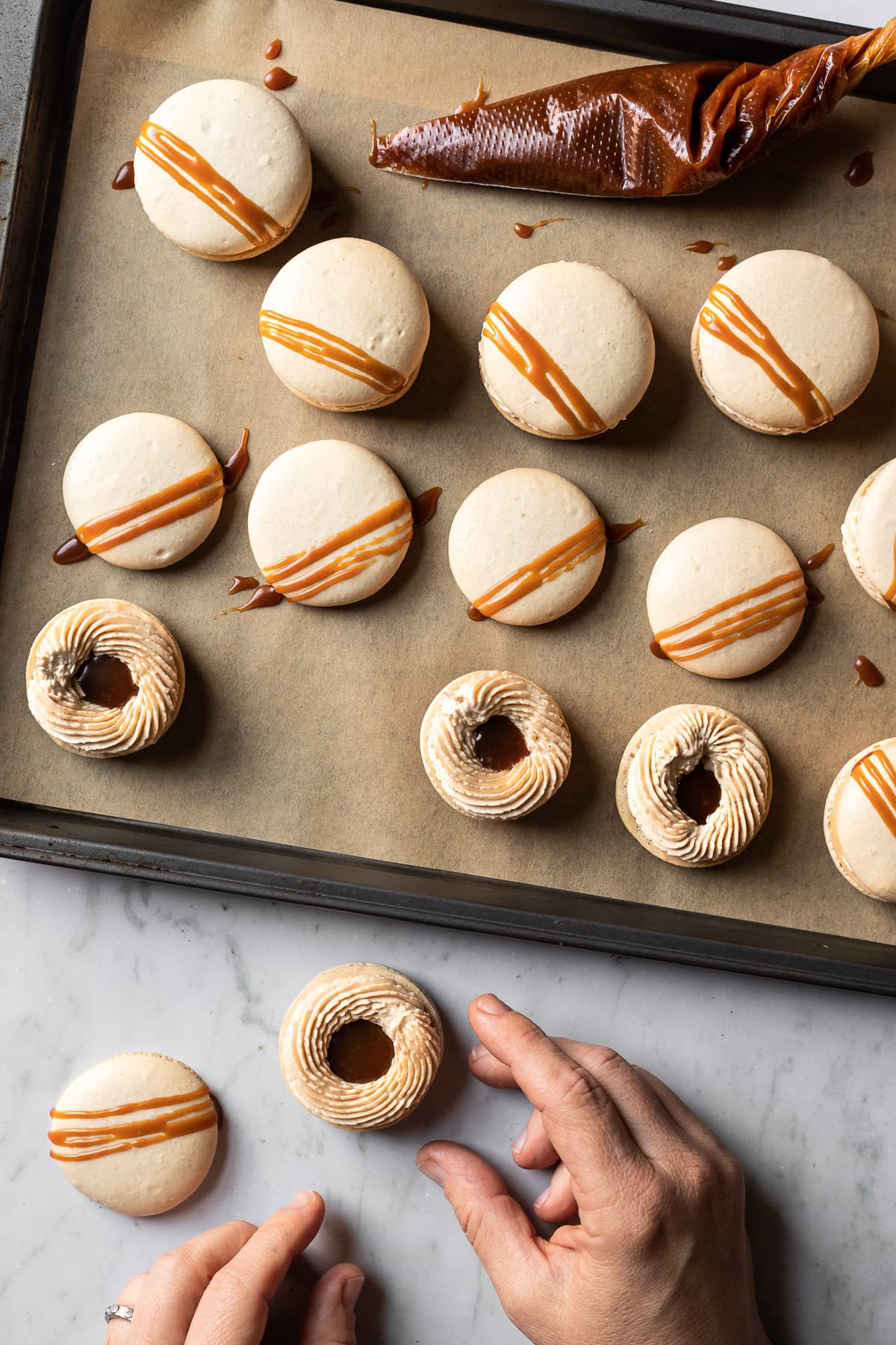 A process photo showing hands reaching into the frame to assemble the top and bottom of macarons. A baking sheet with partially assembled macarons rests nearby.