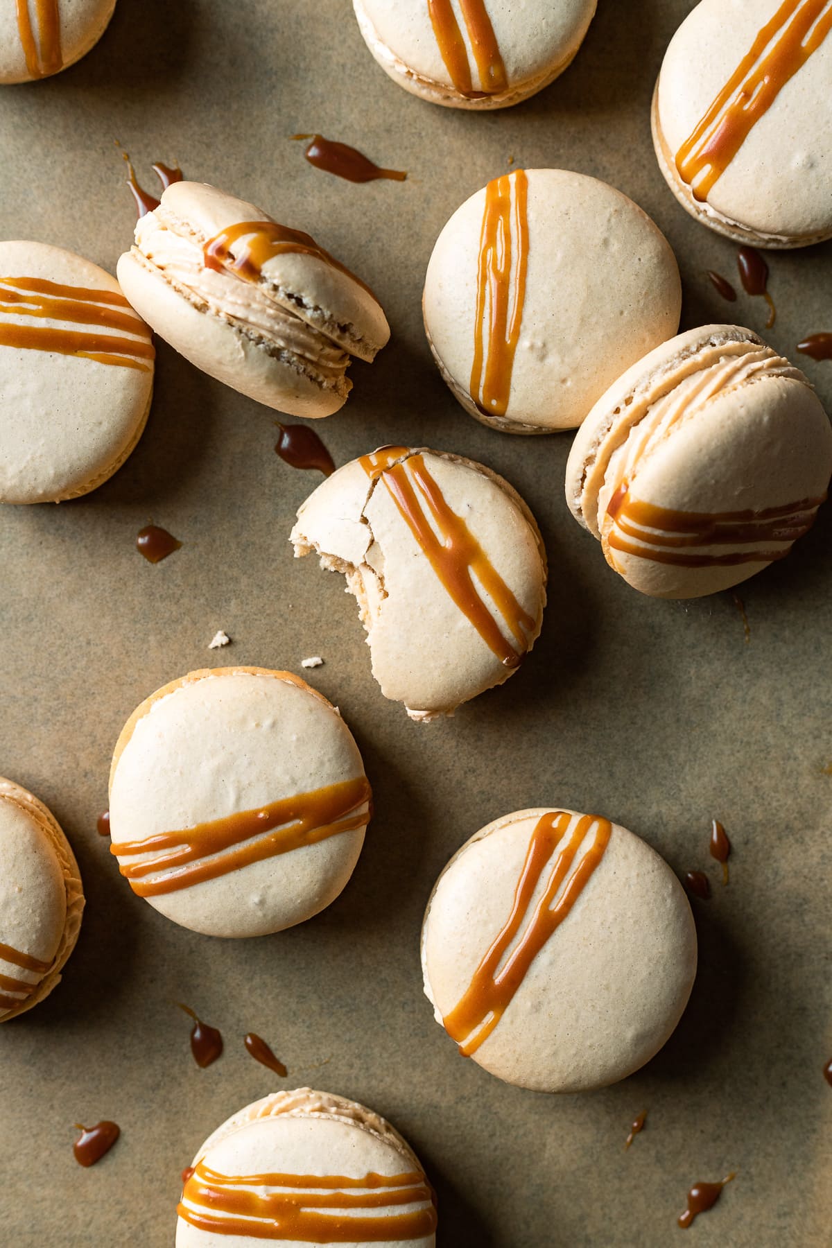 Close up of salted caramel macarons drizzled with caramel sauce on brown parchment surface. There are drips of caramel, and one macaron has a bite out of it.