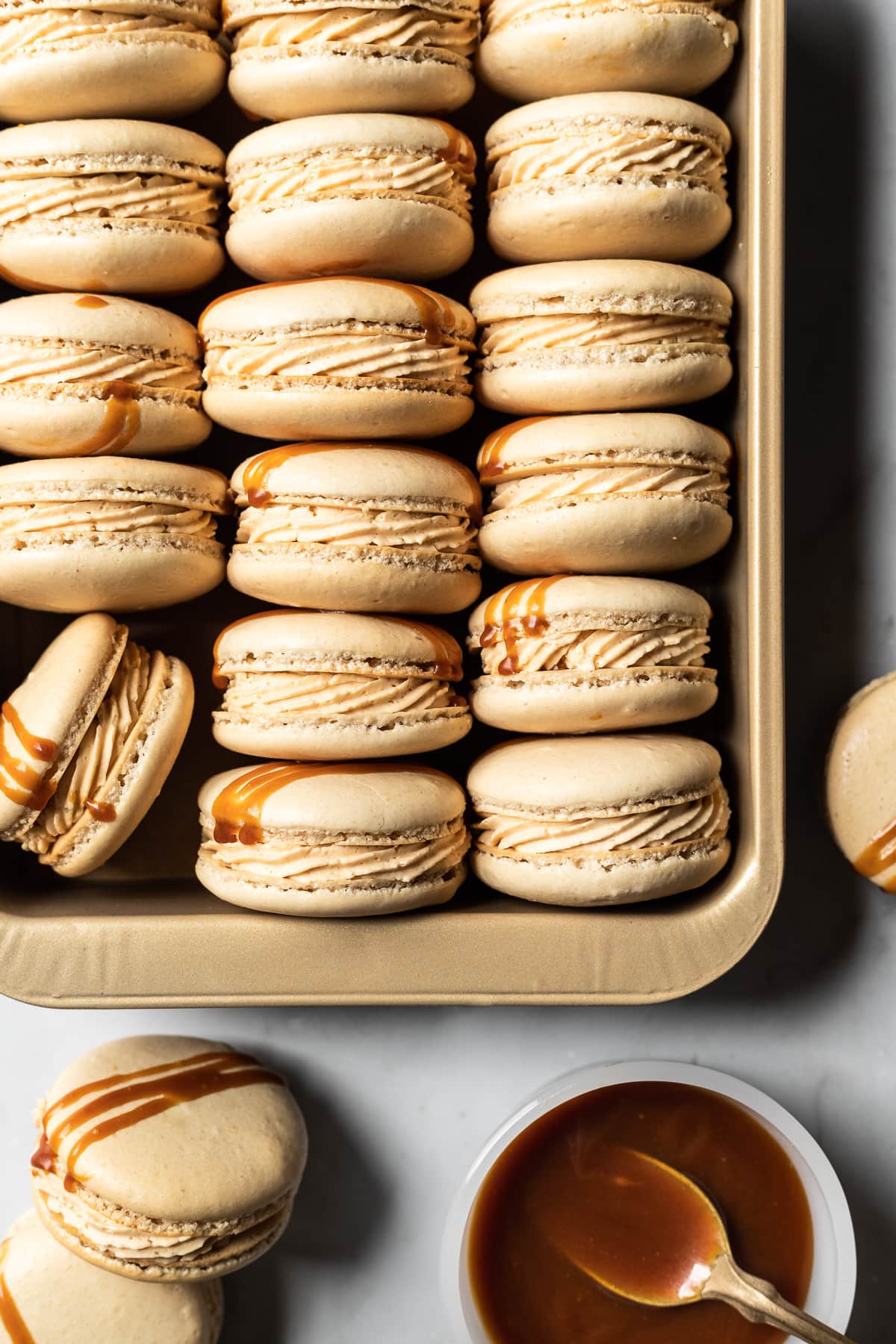 Lightly golden macarons are tucked on their sides into a golden colored baking pan. The decorative piping of the caramel buttercream is visible from the side. A bowl of caramel sauce rests nearby next to additional macarons on a white marble background..