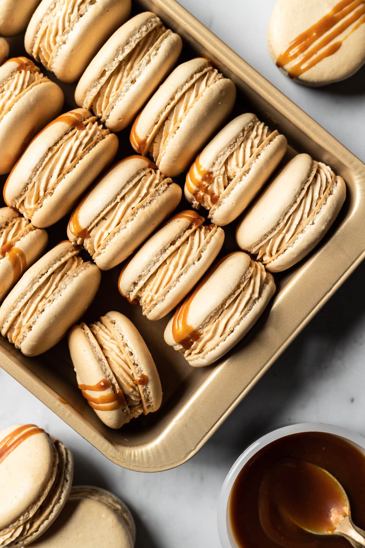 Close up view of salted caramel macarons tipped on their side and nestled into a gold metal pan. The pan is at an angle, and additional macarons and a small bowl of salted caramel peek into the frame.