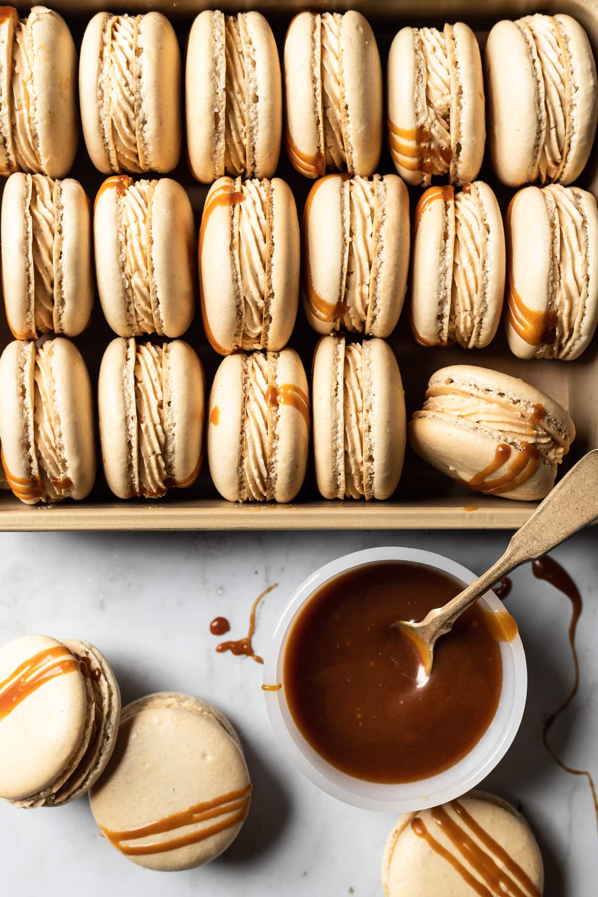 Macarons tipped on their side and nestled into a gold metal pan. A small bowl of salted caramel is at the bottom right of the frame with a vintage gold spoon in it. Caramel drips are on the white marble background surface along with three additional macarons.