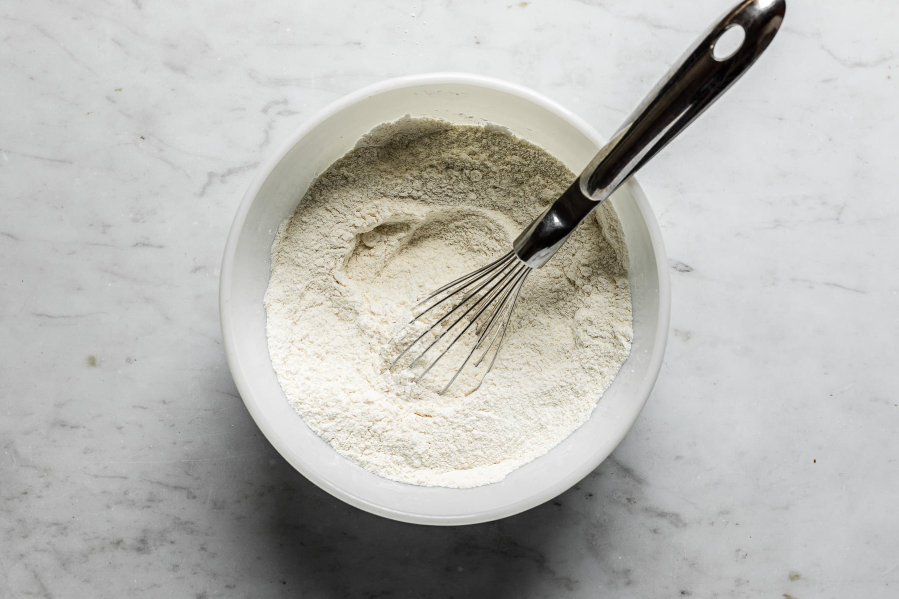 A process image showing dry ingredients in a large metal bowl. It rests on a white marble surface.