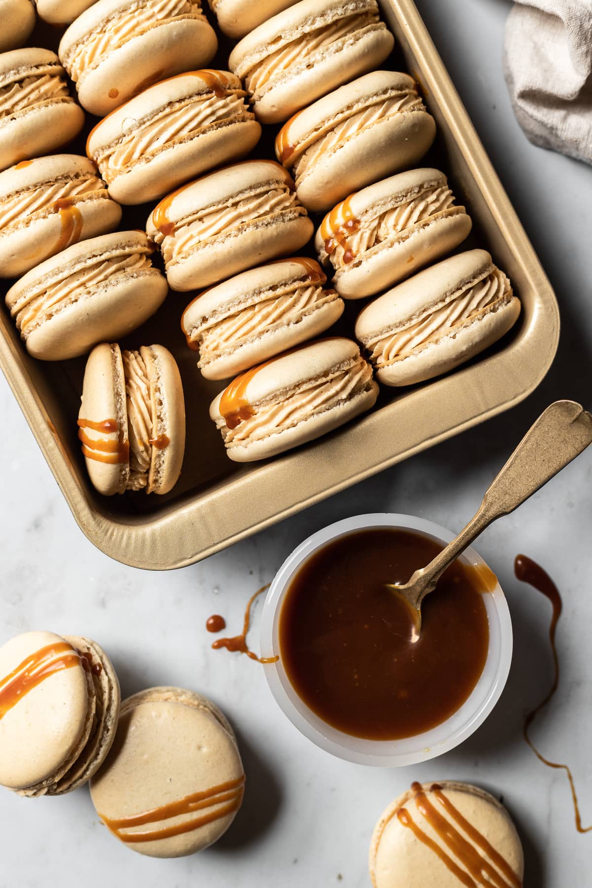 Macarons tipped on their side and nestled into a gold metal pan. A small bowl of salted caramel is at the bottom right of the frame with a vintage gold spoon in it. Caramel drips are on the white marble background surface along with three additional macarons.