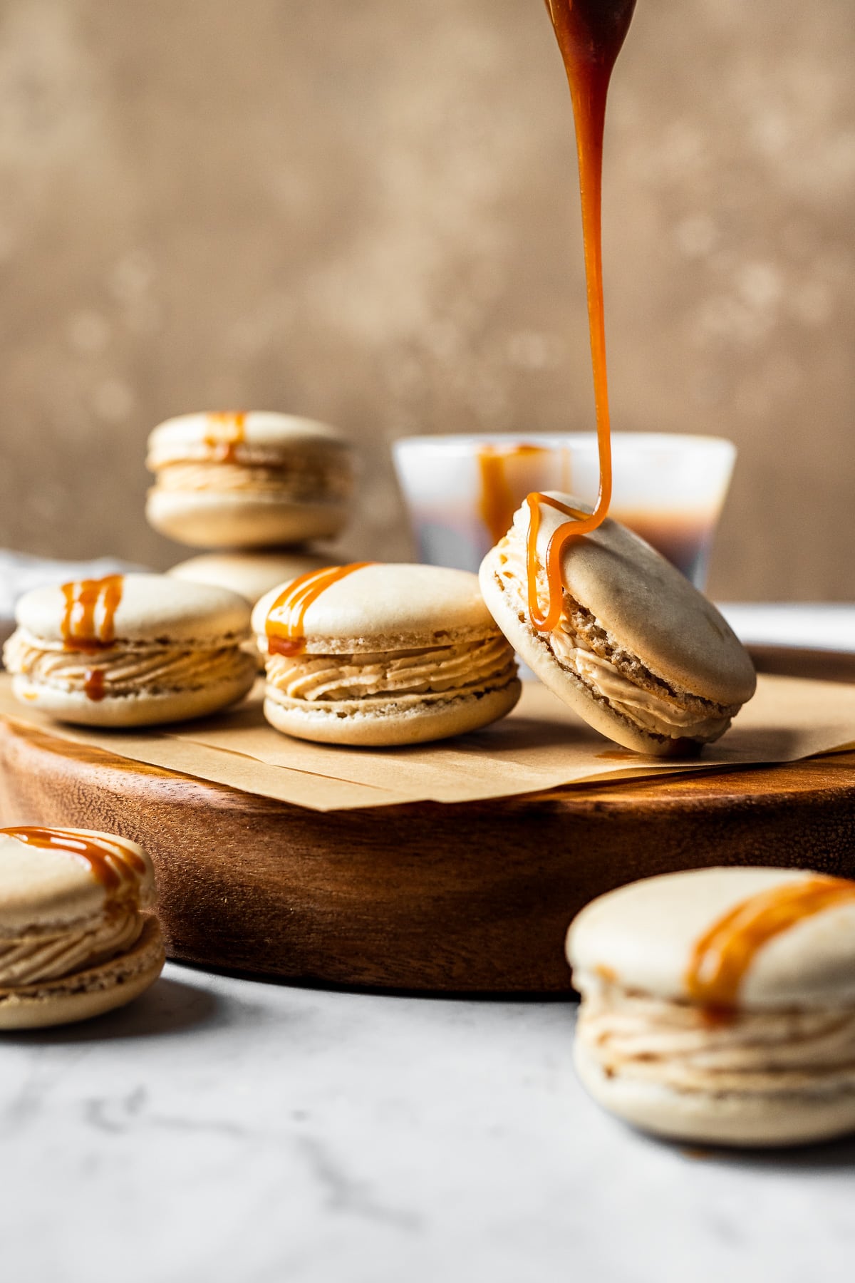 Macarons on a wooden cutting board on a white marble surface with a textured brown background. There is a bowl of caramel sauce out of focus in the background. A drizzle of caramel is pouring from above the frame and dripping onto one of the macarons to create a decorative stripe.