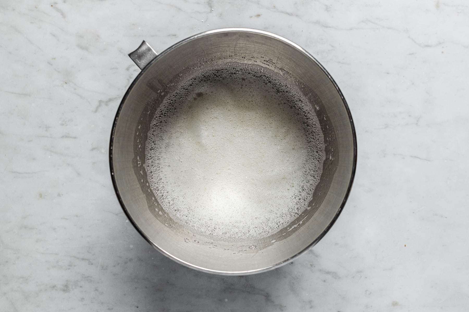 A process image showing foamy egg whites in a silver metal mixing bowl on a white marble surface.