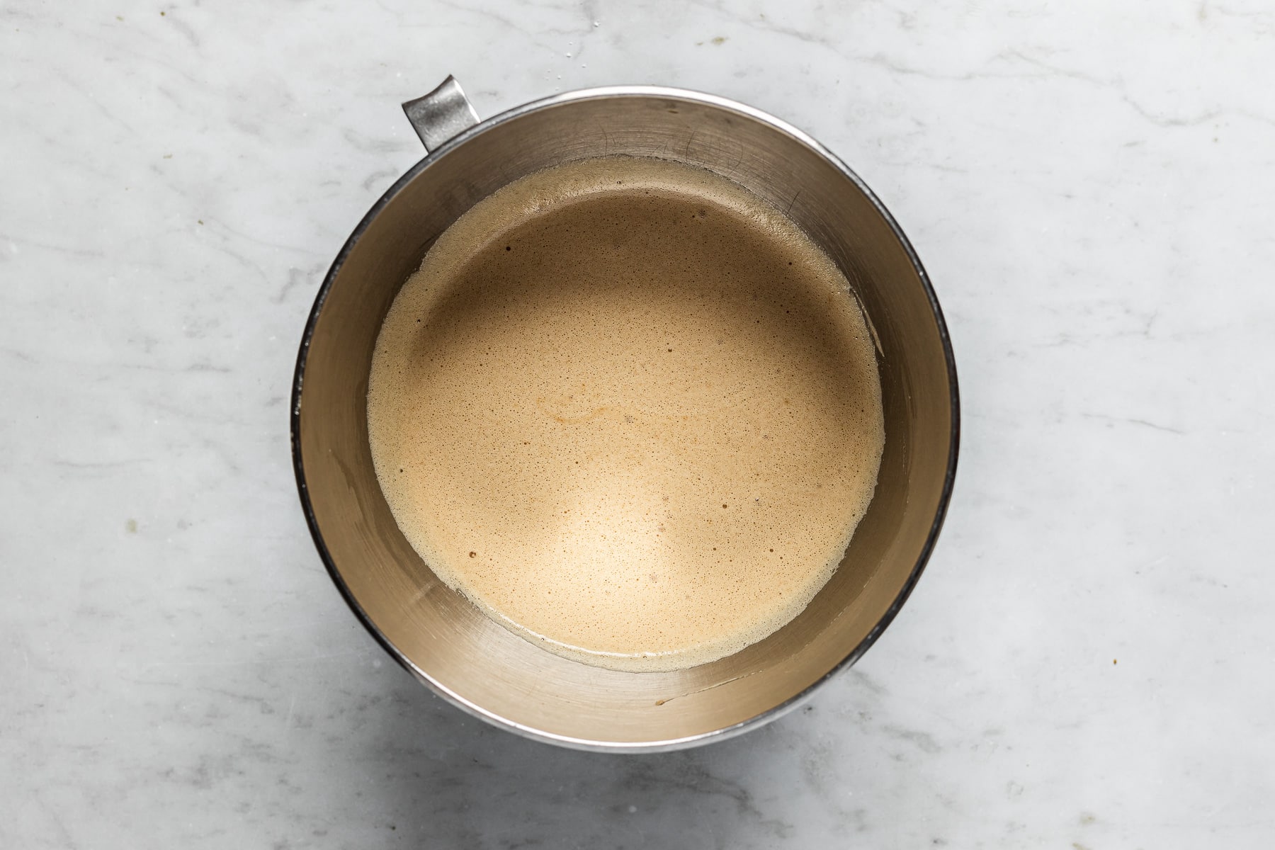 A process photo showing a silver mixing bowl with egg whites and brown sugar mixed together. The bowl rests on a white marble surface.