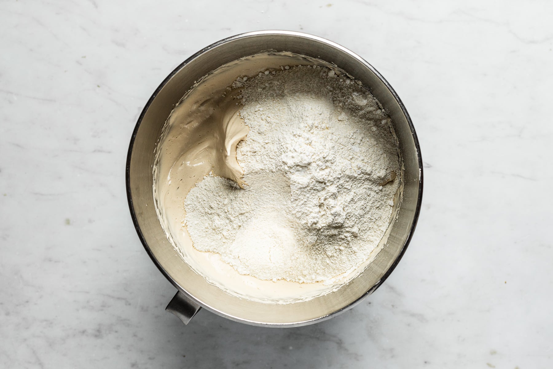 A process image showing dry ingredients mixed into stiffly beaten eggwhites in a large metal bowl. It rests on a white marble surface.