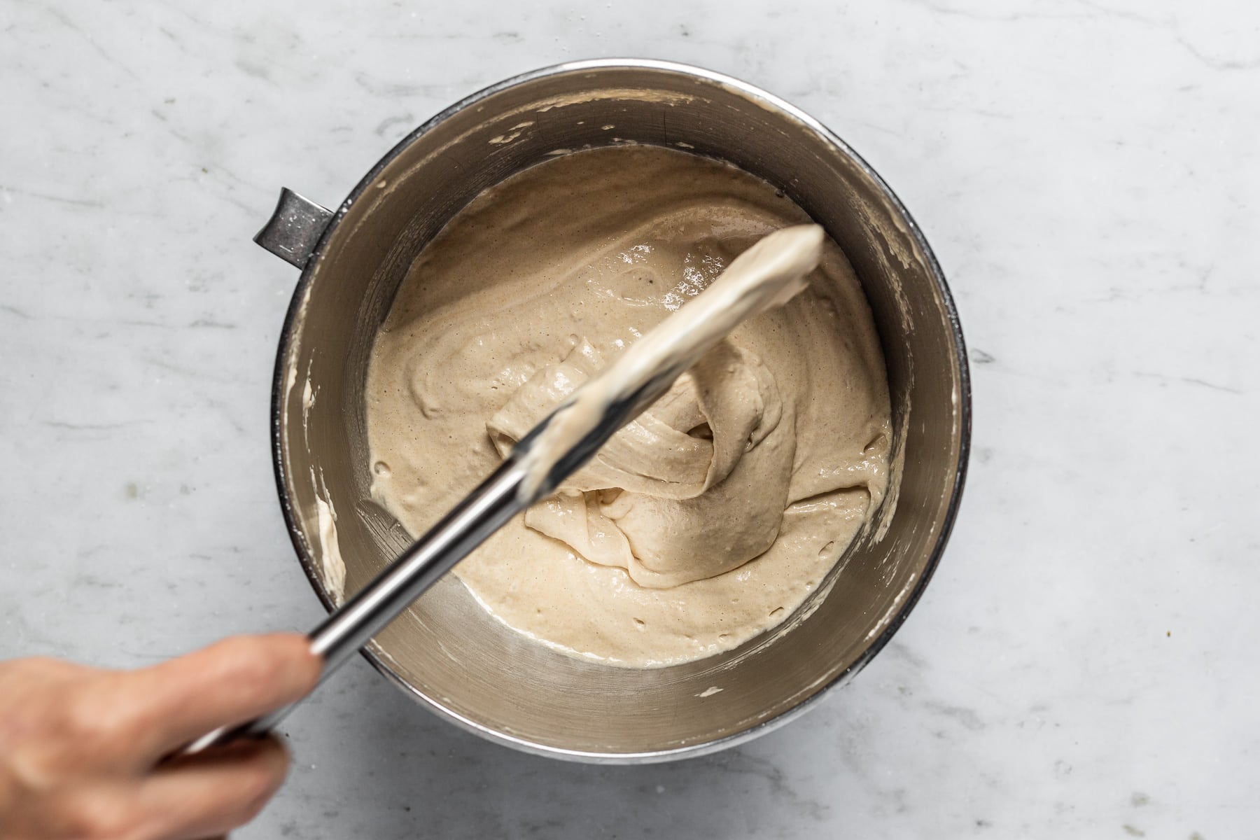 Process shot showing macaron batter dripping off the spatula in a slow figure eight shape, demonstrating batter is ready. Batter is in a metal bowl on a white marble surface and a hand holds the spatula above the bowl.