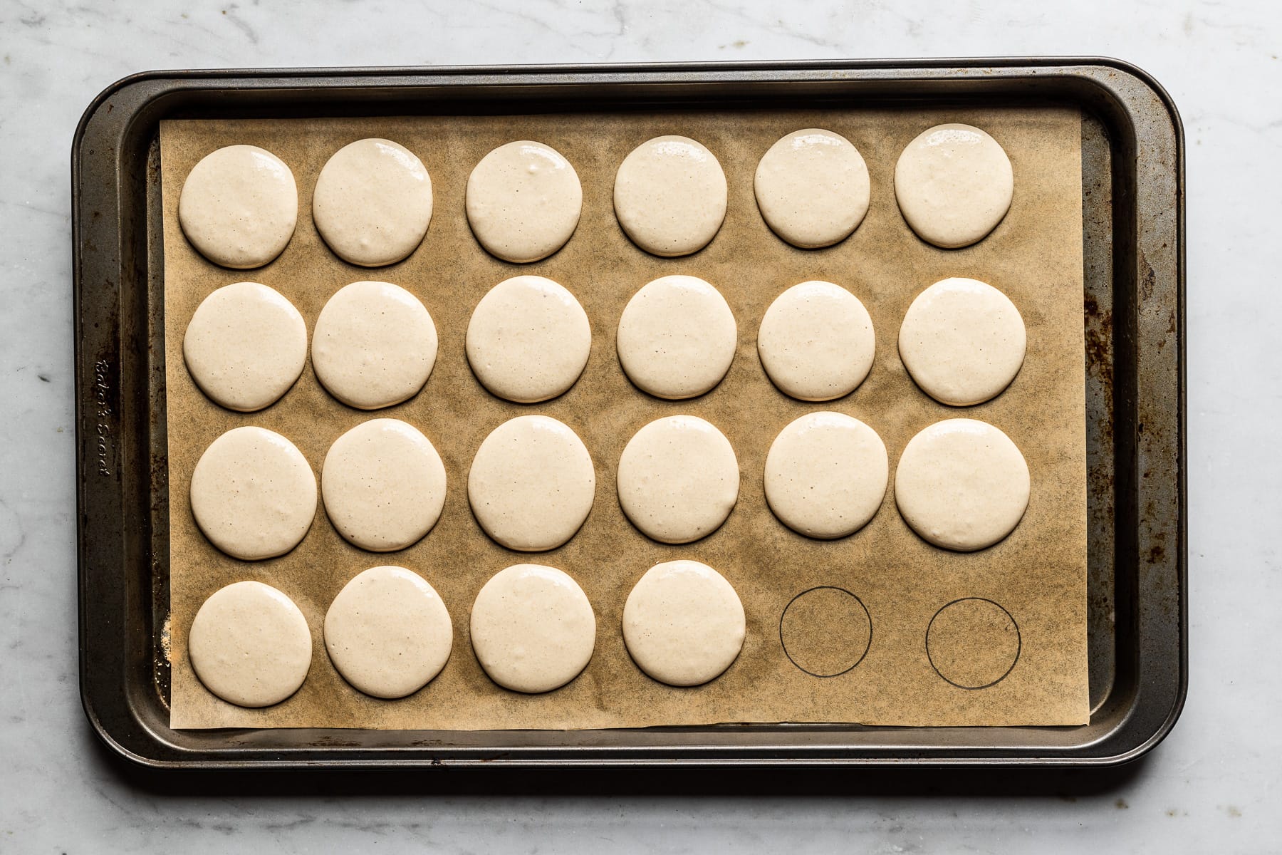 Process photo of macaron batter piped into circles on a parchment lined baking sheet. The pan rests on a white marble surface.