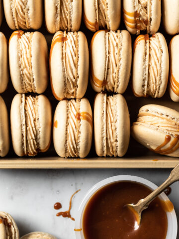 Close up view of salted caramel macarons tipped on their side and nestled into a gold metal pan. A small bowl of salted caramel is at the bottom right of the frame with a vintage gold spoon in it. Caramel drips are on the white marble background surface.
