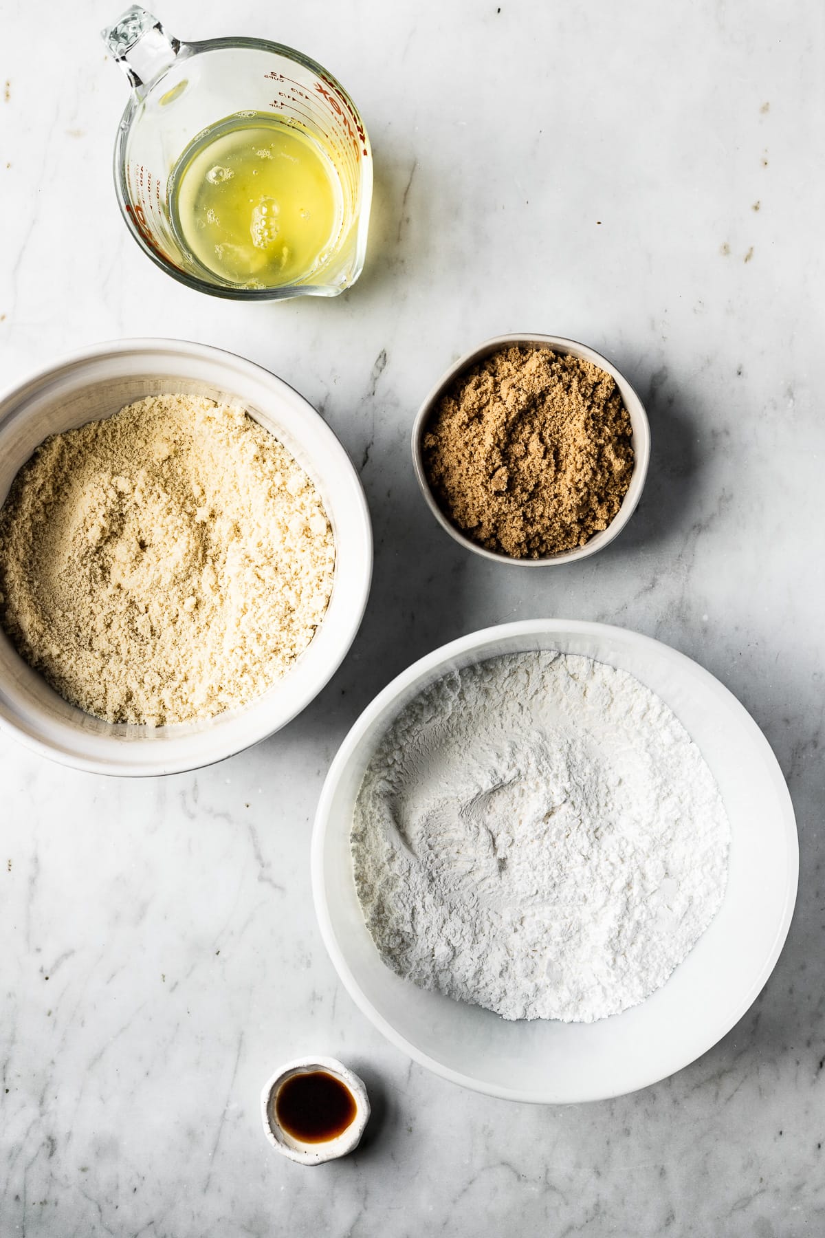 Ingredients for macarons in white ceramic bowls on a white marble surface. Ingredients include egg whites, brown sugar, almond flour, powdered sugar and vanilla extract.