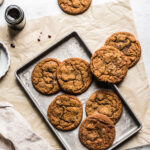 Ginger cookies on a baking sheet resting on a brown parchment paper rectangle. A jar of molasses is nearby.