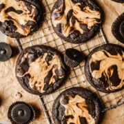 Cooled dark chocolate peanut butter cookies on a vintage metal cooling rack on top of brown parchment paper. Peanut butter cups in dark brown paper are scattered nearby.