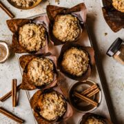 Streusel topped muffins in brown paper parchment liners in a muffin tin surrounded by cinnamon sugar and cinnamon sticks.