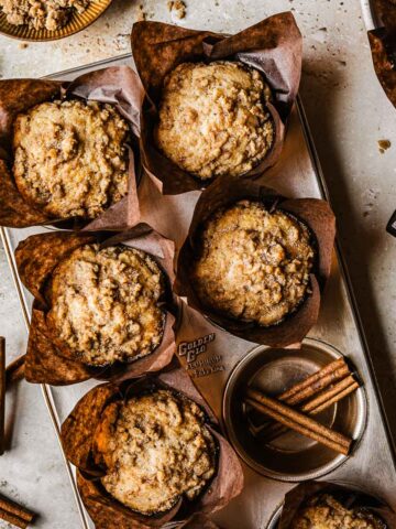 Streusel topped muffins in brown paper parchment liners in a muffin tin surrounded by cinnamon sugar and cinnamon sticks.