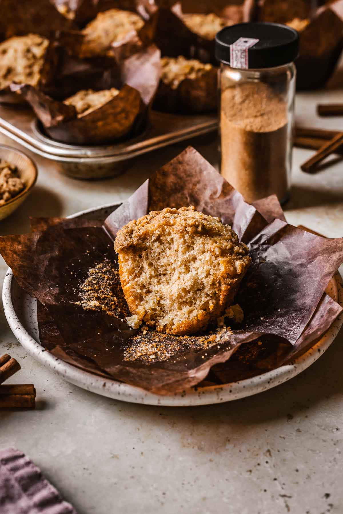 A cinnamon streusel muffin on brown paper parchment squares on a white ceramic plate. In the background is a glass jar of cinnamon sugar and a full tin of baked muffins.