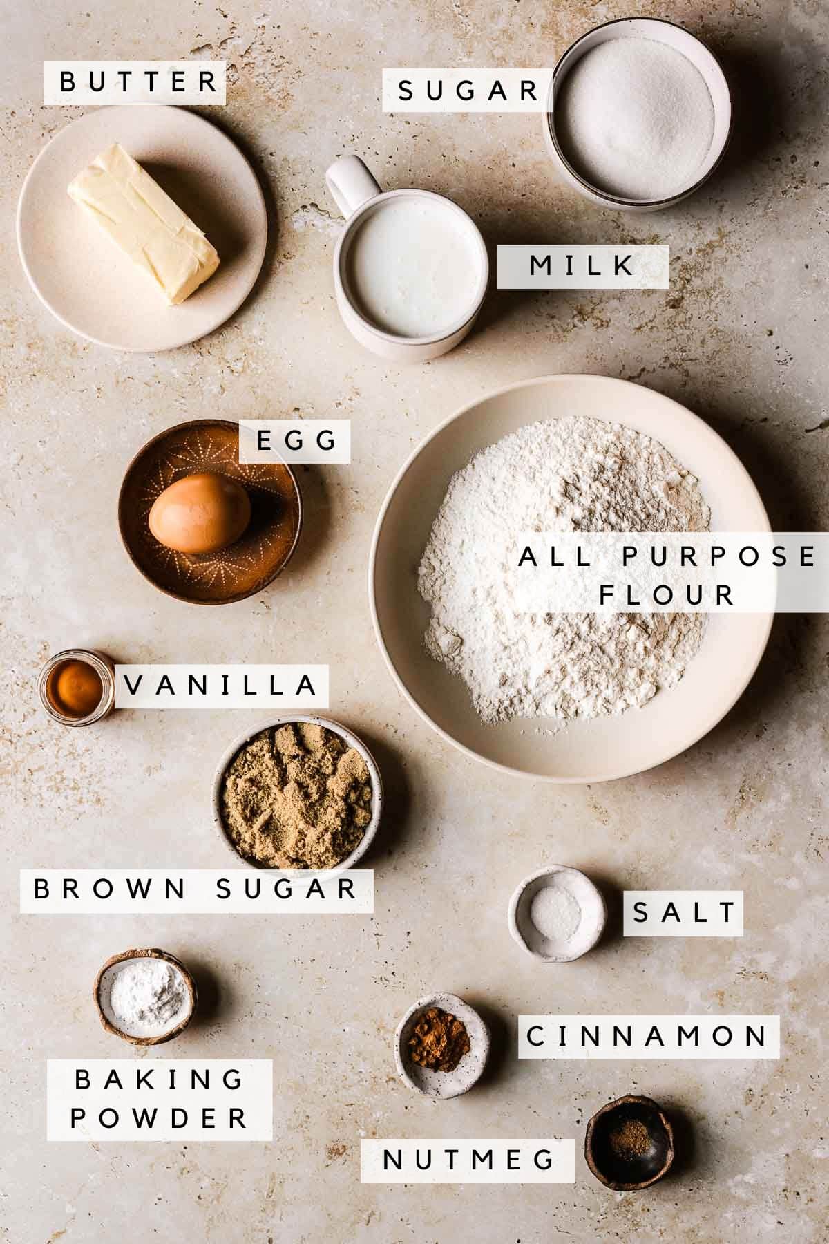 Small bowls with ingredients for cinnamon streusel muffins on a tan stone background. There are labels identifying each ingredient.