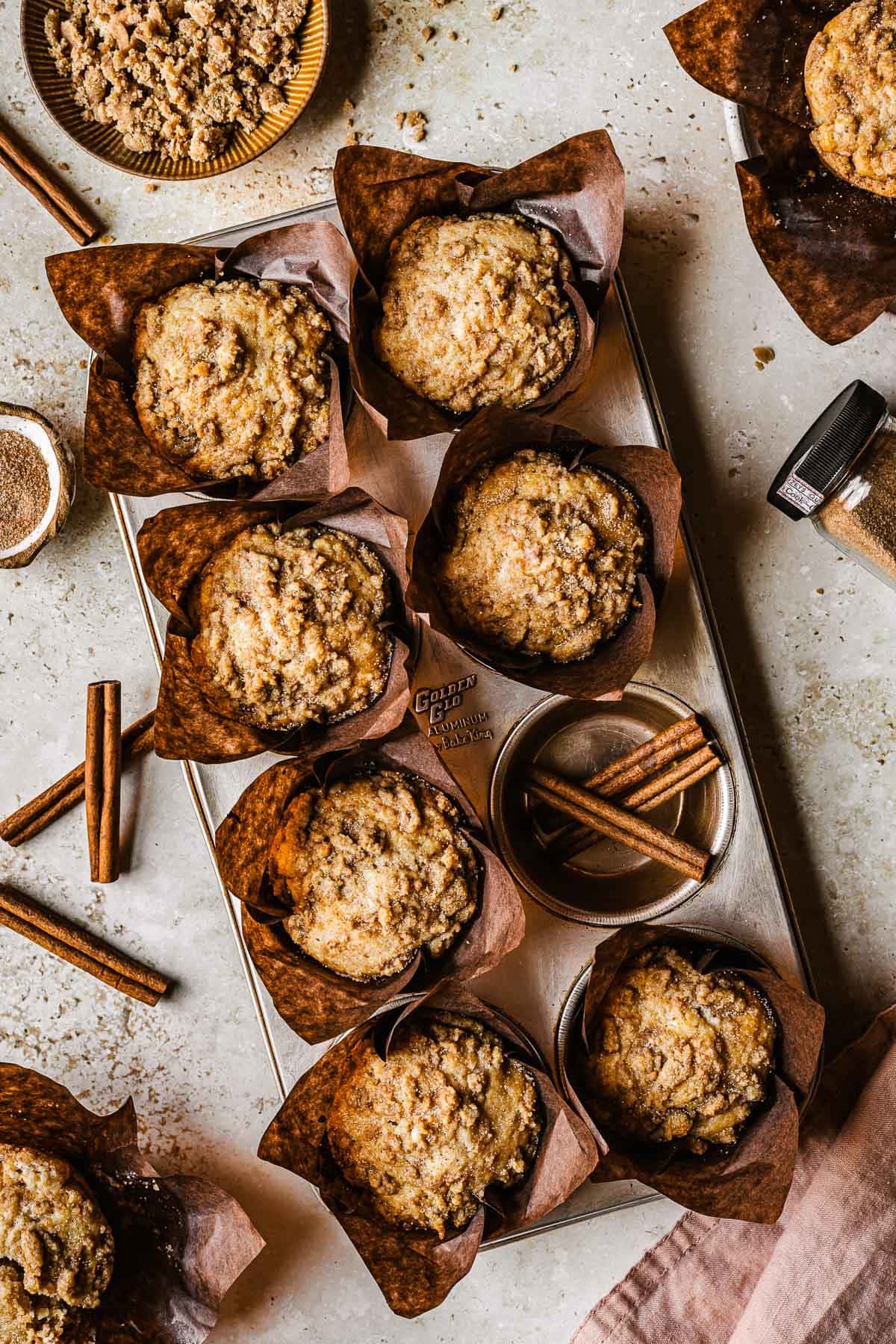 Streusel topped muffins in brown paper parchment liners in a muffin tin surrounded by cinnamon sugar, cinnamon sticks, a bowl of streusel and a pink napkin.