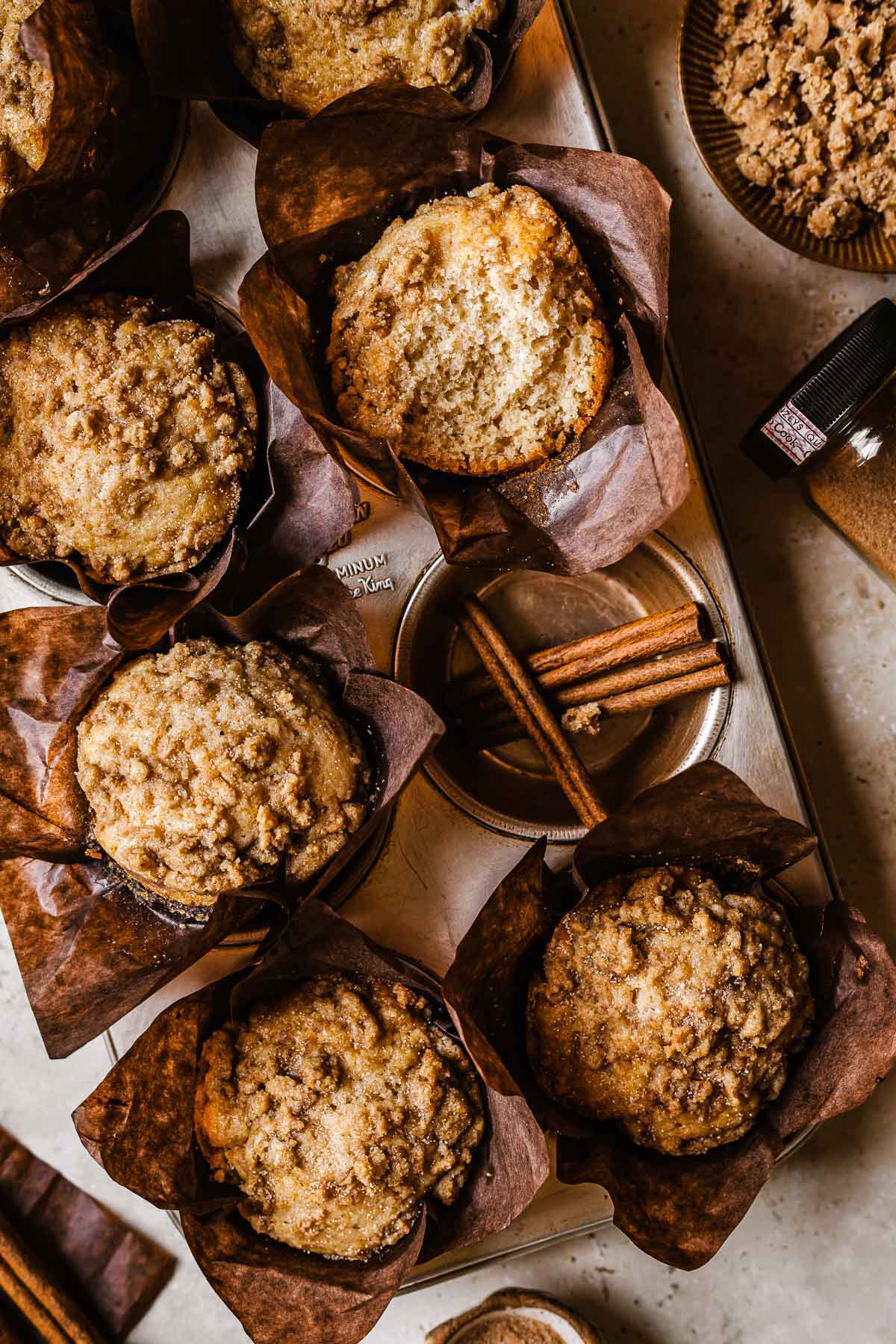 Cinnamon streusel muffins in brown parchment paper wrappers in a muffin tin. One muffin has been broken open to show the crumb inside. Three cinnamon sticks rest in an empty muffin tin cavity.