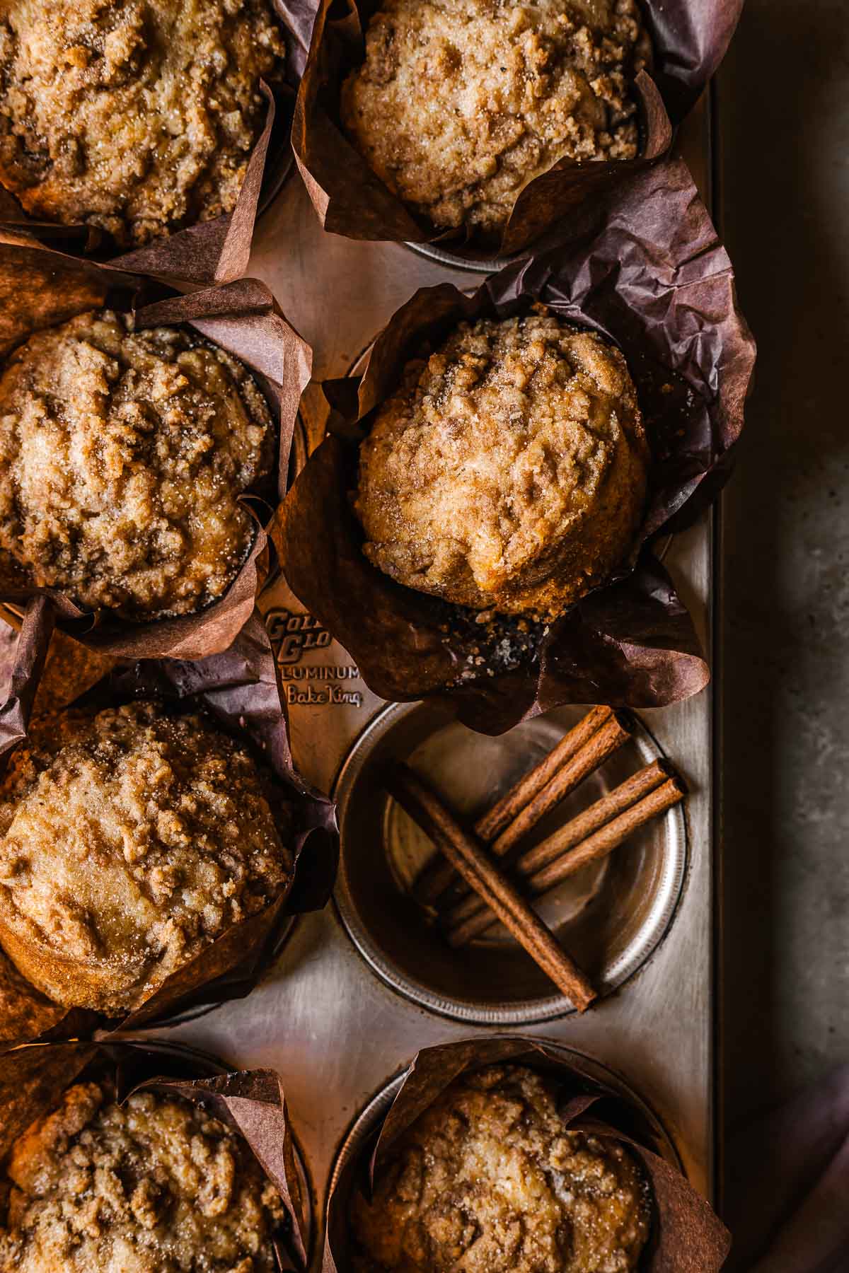 Streusel topped muffins in brown parchment wrappers in a muffin tin. One hole of the muffin tin has three sticks of cinnamon inside.