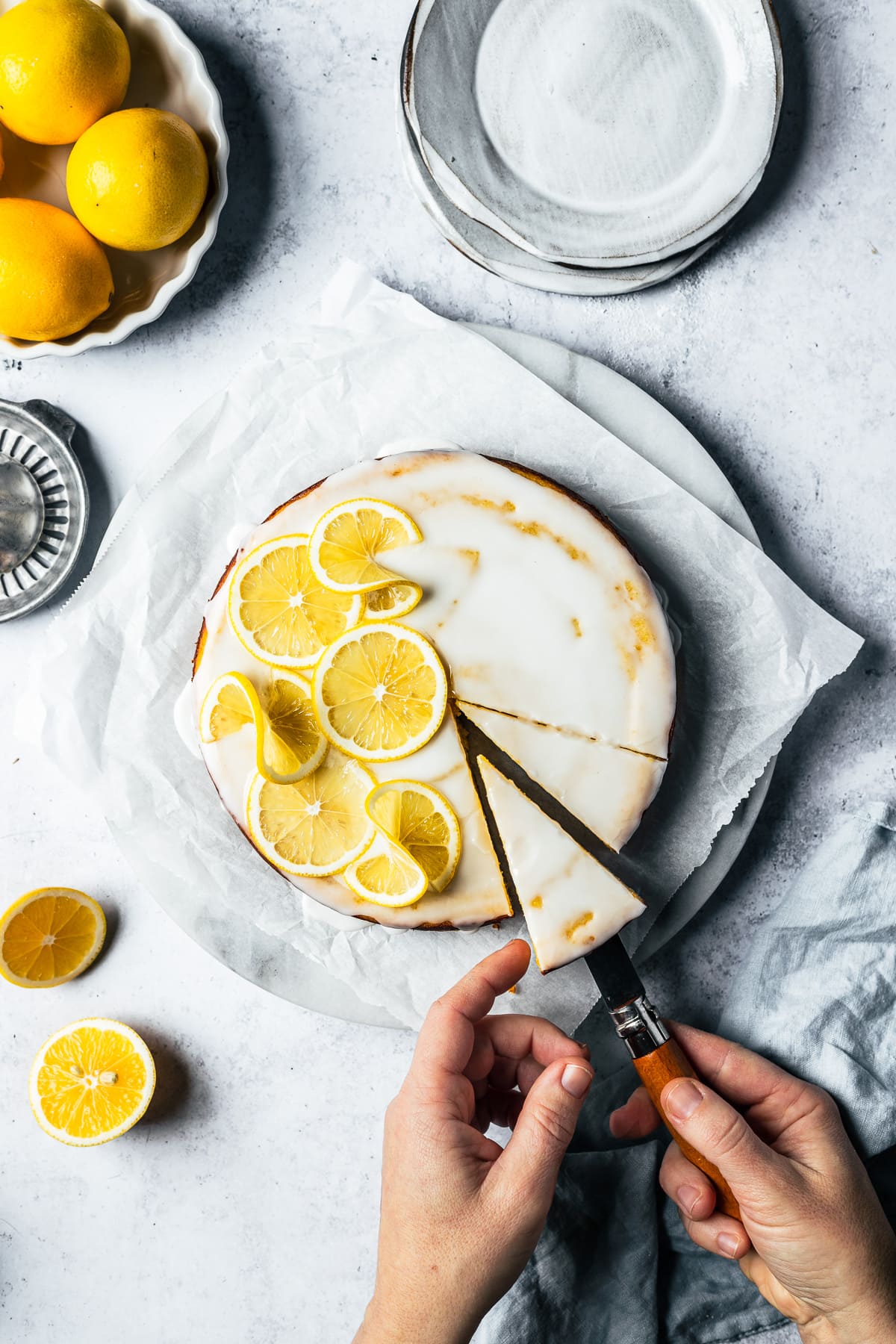 Hands lift a slice of lemon cake from a marble platter. Ceramic plates, lemons, and a juicer surround the cake on a light blue concrete background.