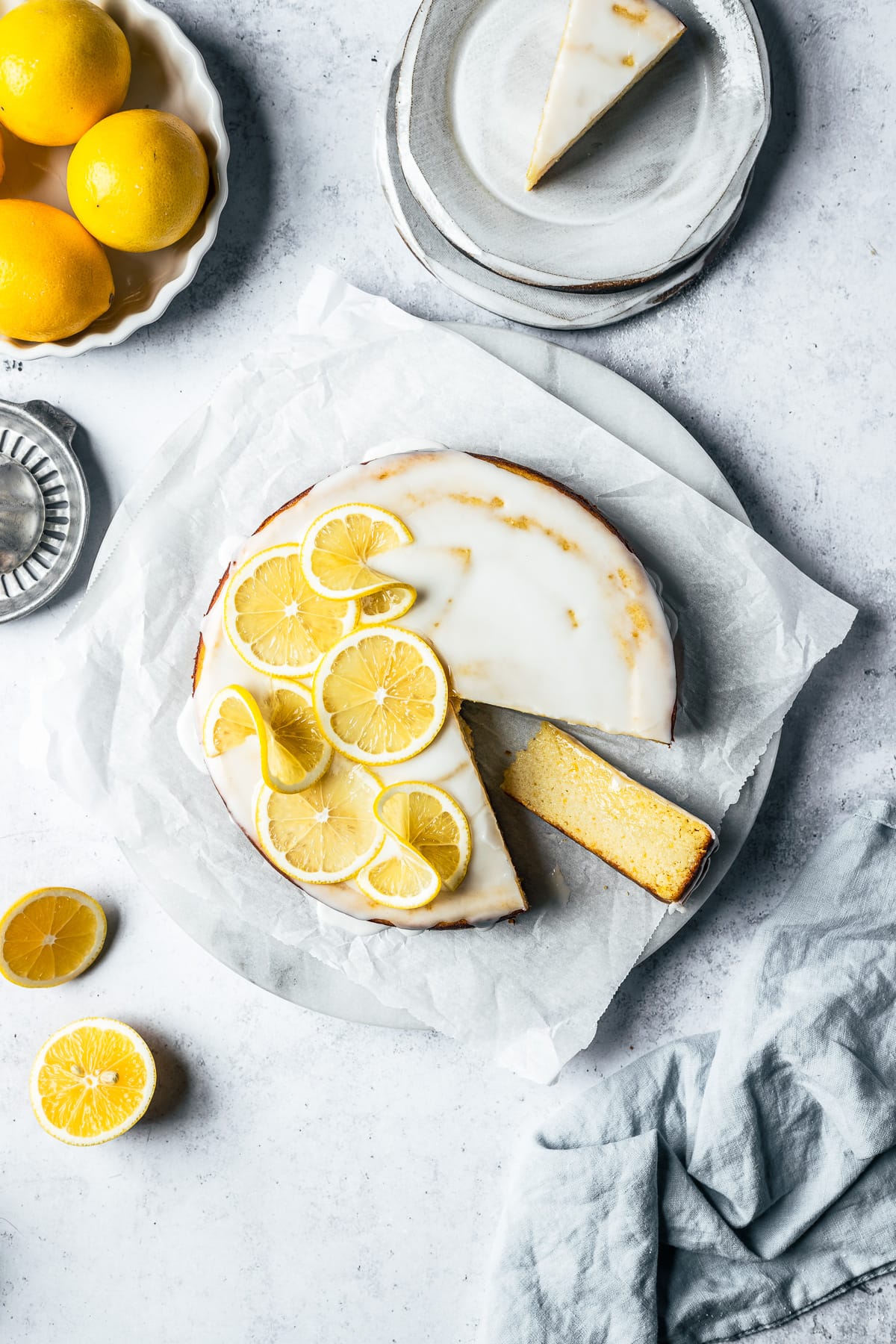 A slice of lemon cake rests on its side next to the rest of the glazed cake decorated with lemon slices. It rests on a marble platter. Ceramic plates, lemons, and a juicer surround the cake on a light blue concrete background.